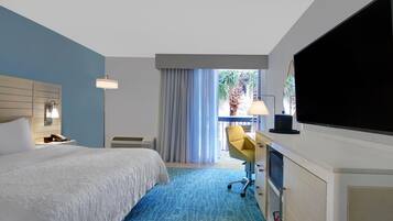 Habitación de hotel con cama, escritorio, silla, televisión y balcón con cortinas.