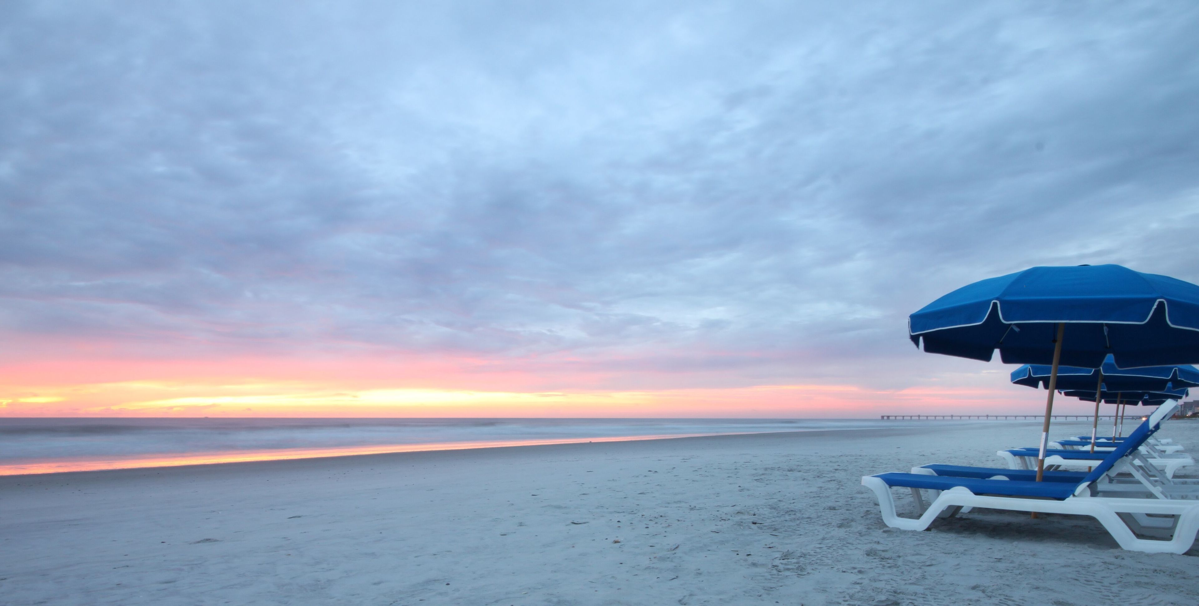 Een strand met een rij blauwe en witte ligbedden en een blauwe parasol.