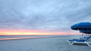 Een strand met een rij blauwe en witte ligbedden en een blauwe parasol.