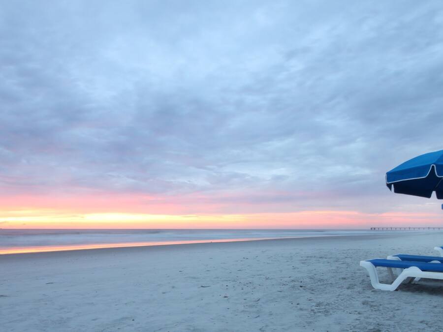 Een strand met een rij blauwe en witte ligbedden en een blauwe parasol.