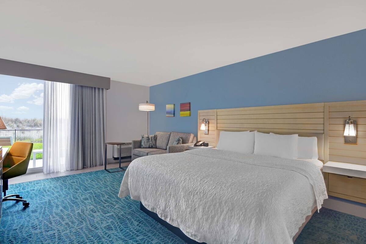 A hotel room with a large bed, a desk, a chair, a small table, and a balcony with curtains.
