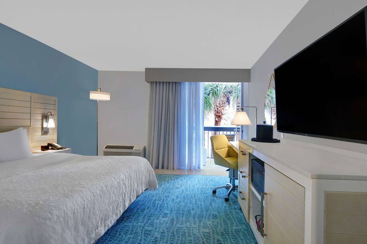 A hotel room with a bed, desk, TV, and a view of palm trees through the window.