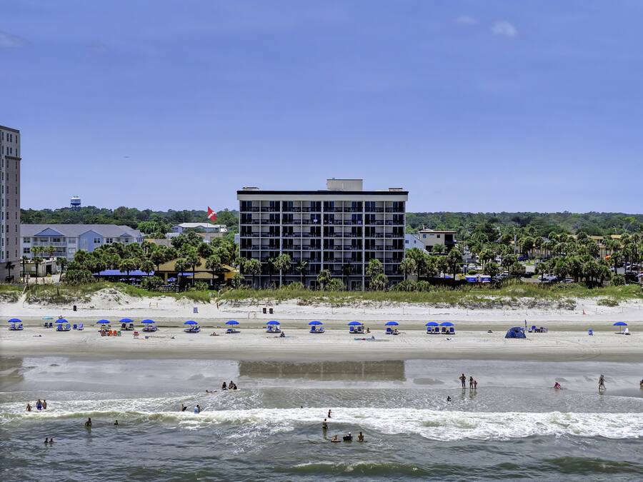 Un hotel frente a la playa con varios pisos, una playa arenosa con sombrillas y vistas al océano.