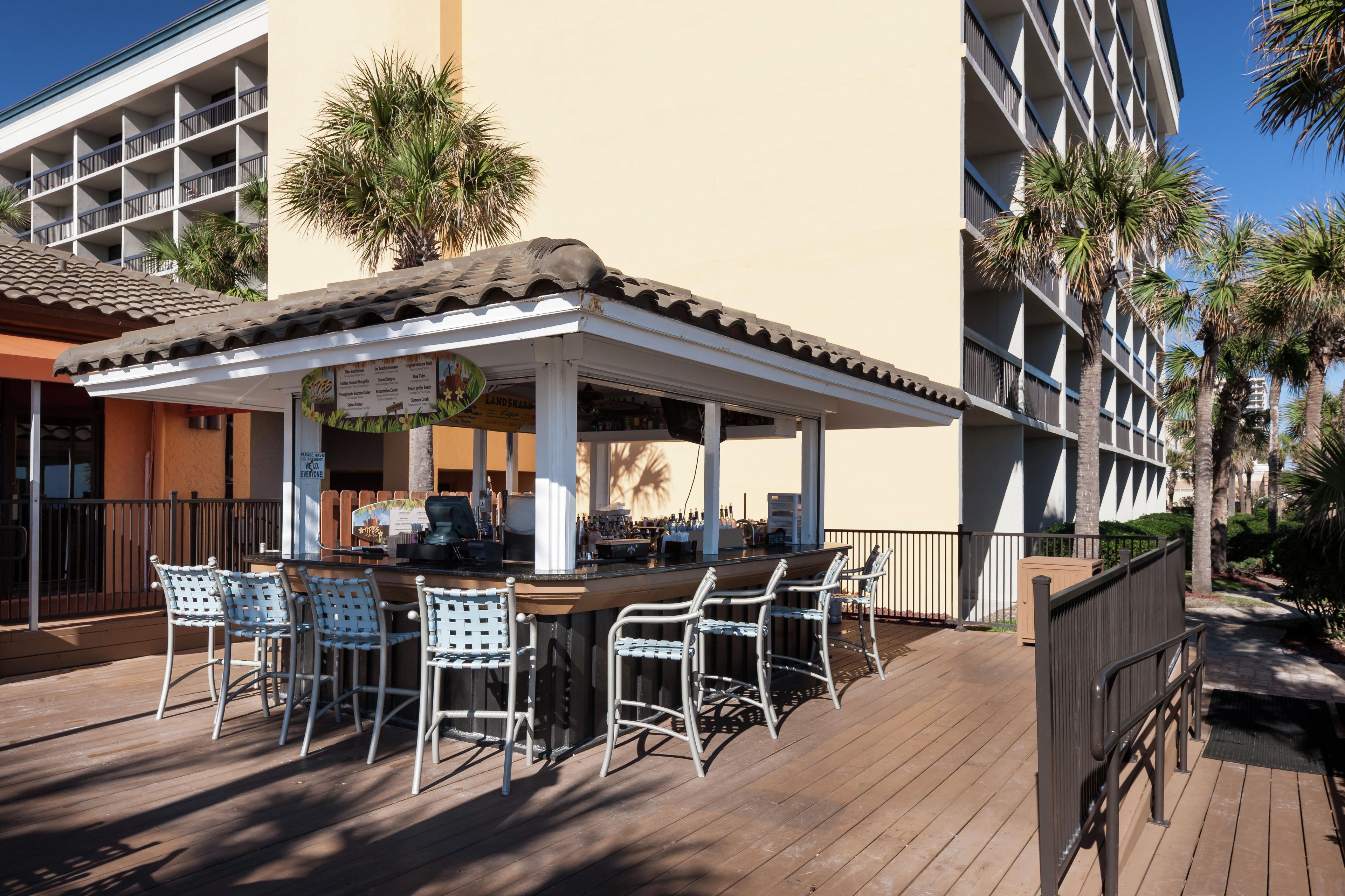 Zona de bar con asientos al aire libre, una sección cubierta y un edificio con balcones y palmeras.