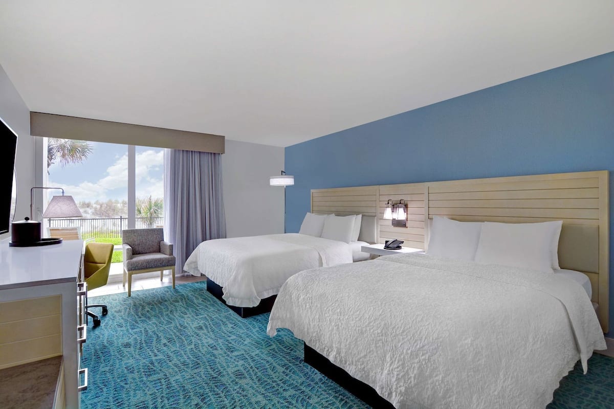 A hotel room with two beds, a desk with a lamp, a chair, and a view of the outdoors.