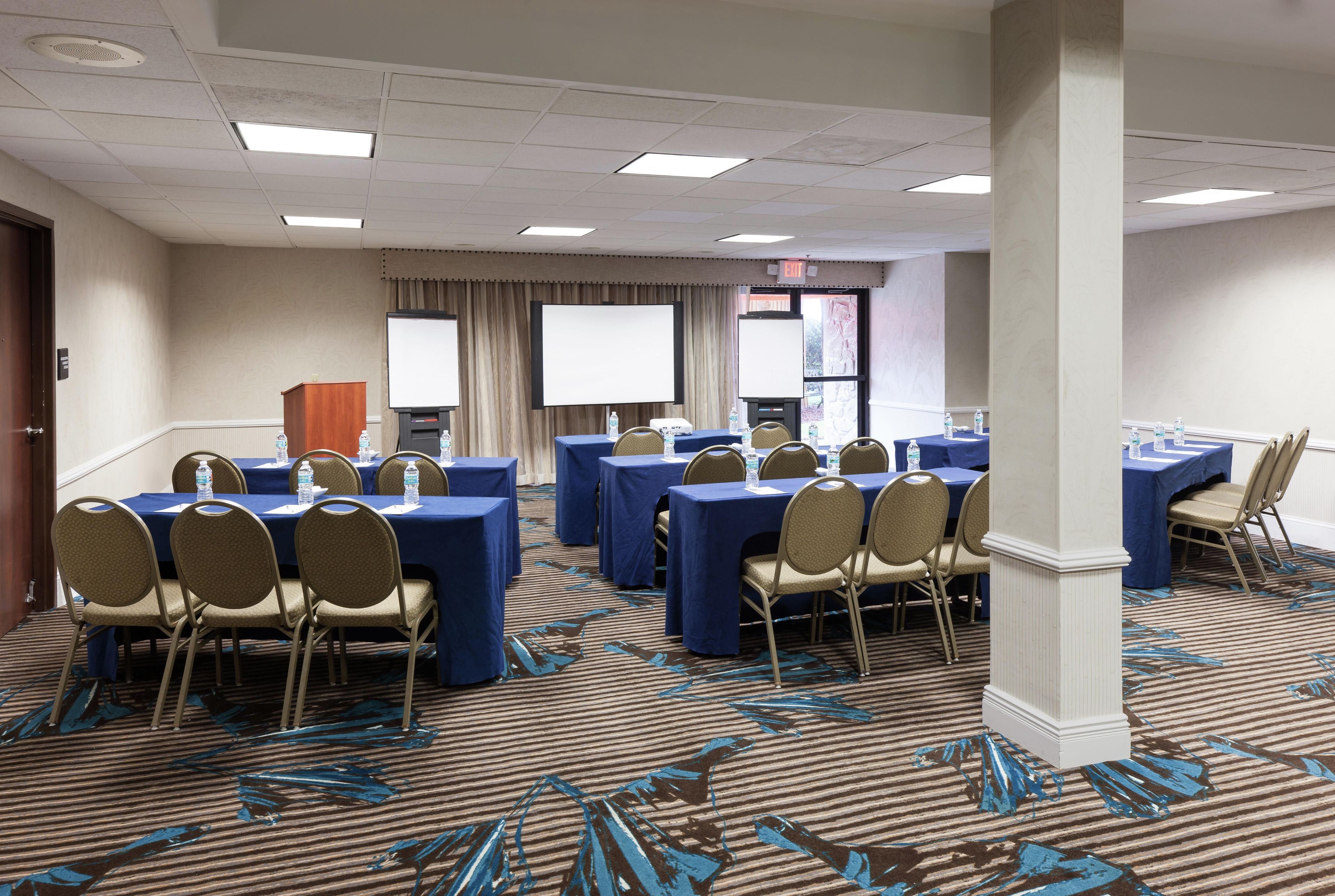 A conference room with tables, chairs, a projector screen, and a carpeted floor.
