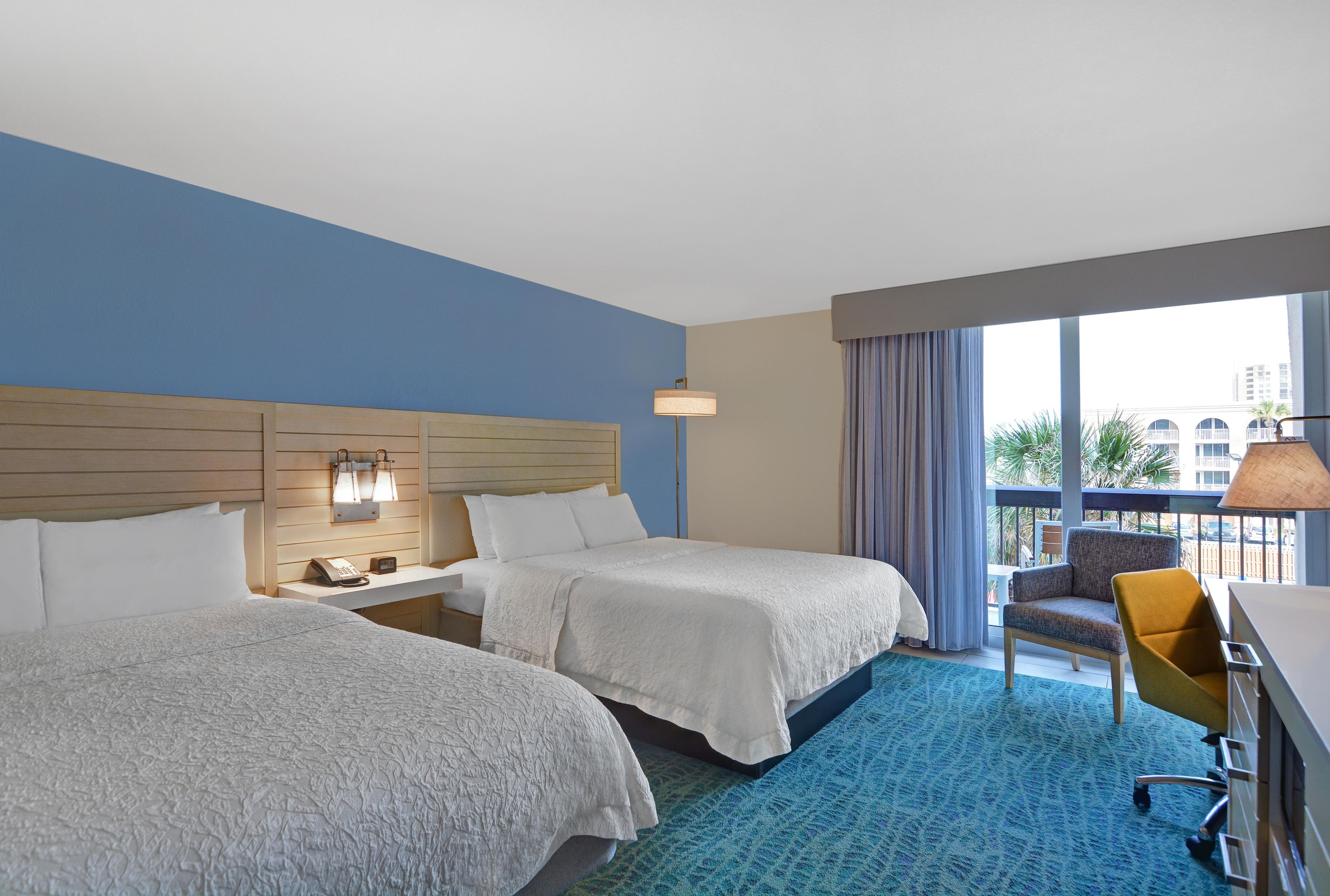 A hotel room with two beds, a desk, a chair, and a balcony with a view.