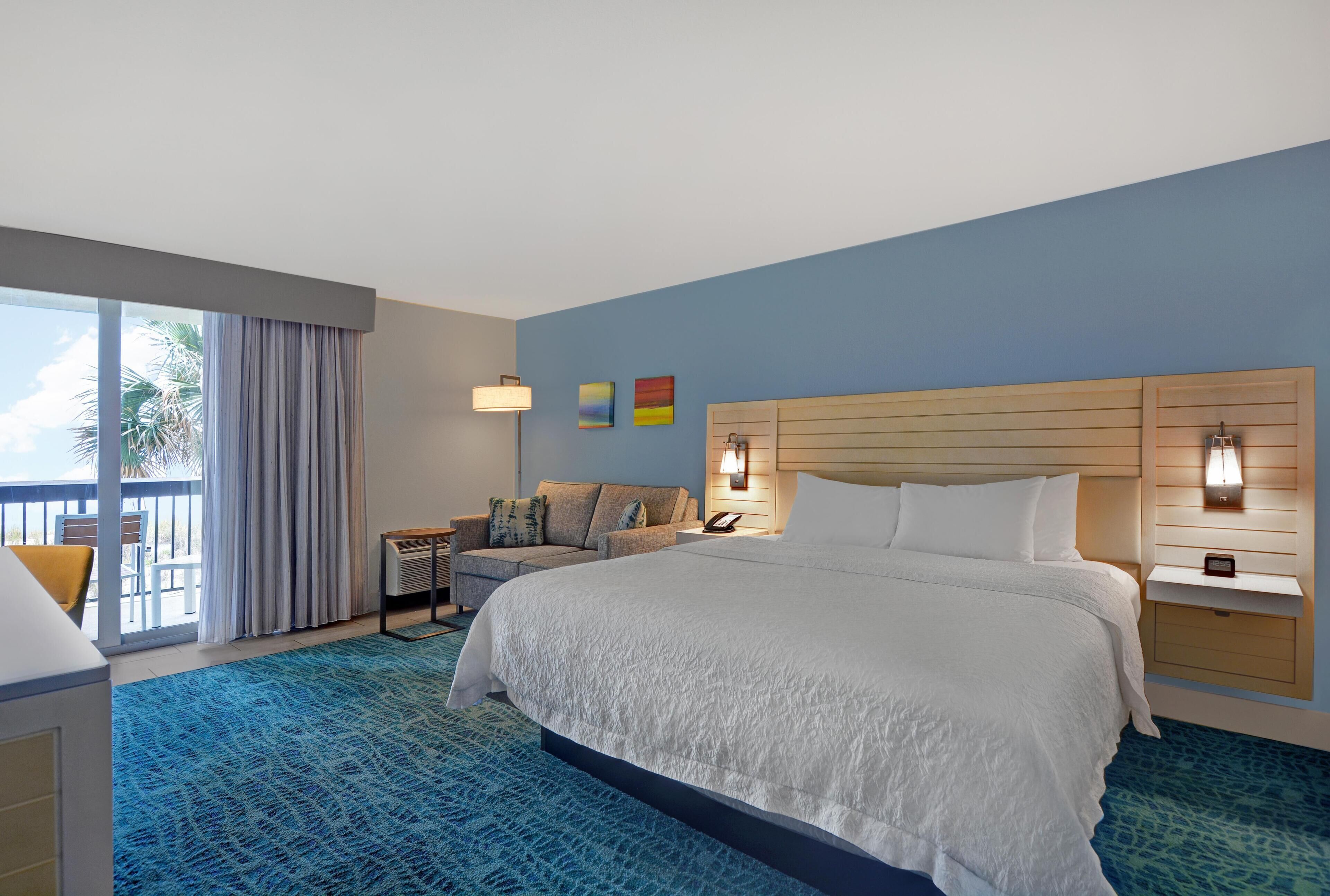 A hotel room with a large bed, a desk, a chair, a nightstand, and a view of the outdoors.