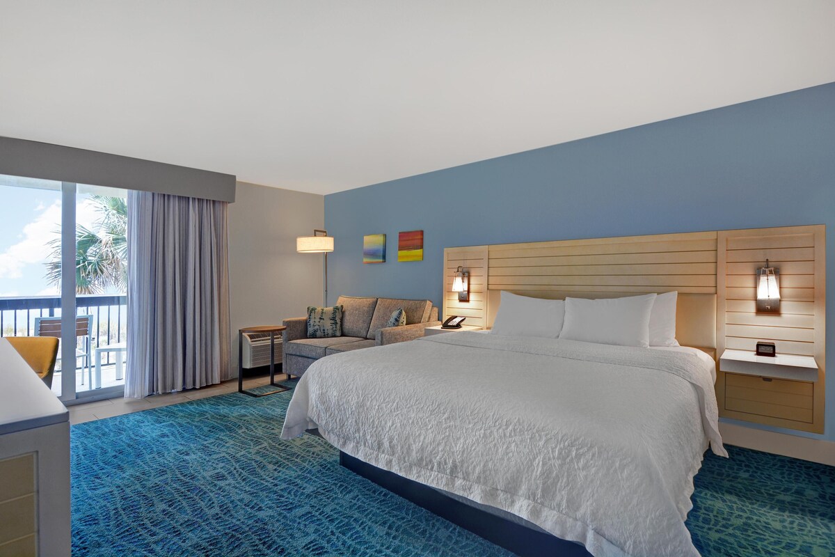 A hotel room with a large bed, a desk, a chair, a nightstand, and a view of the outdoors.