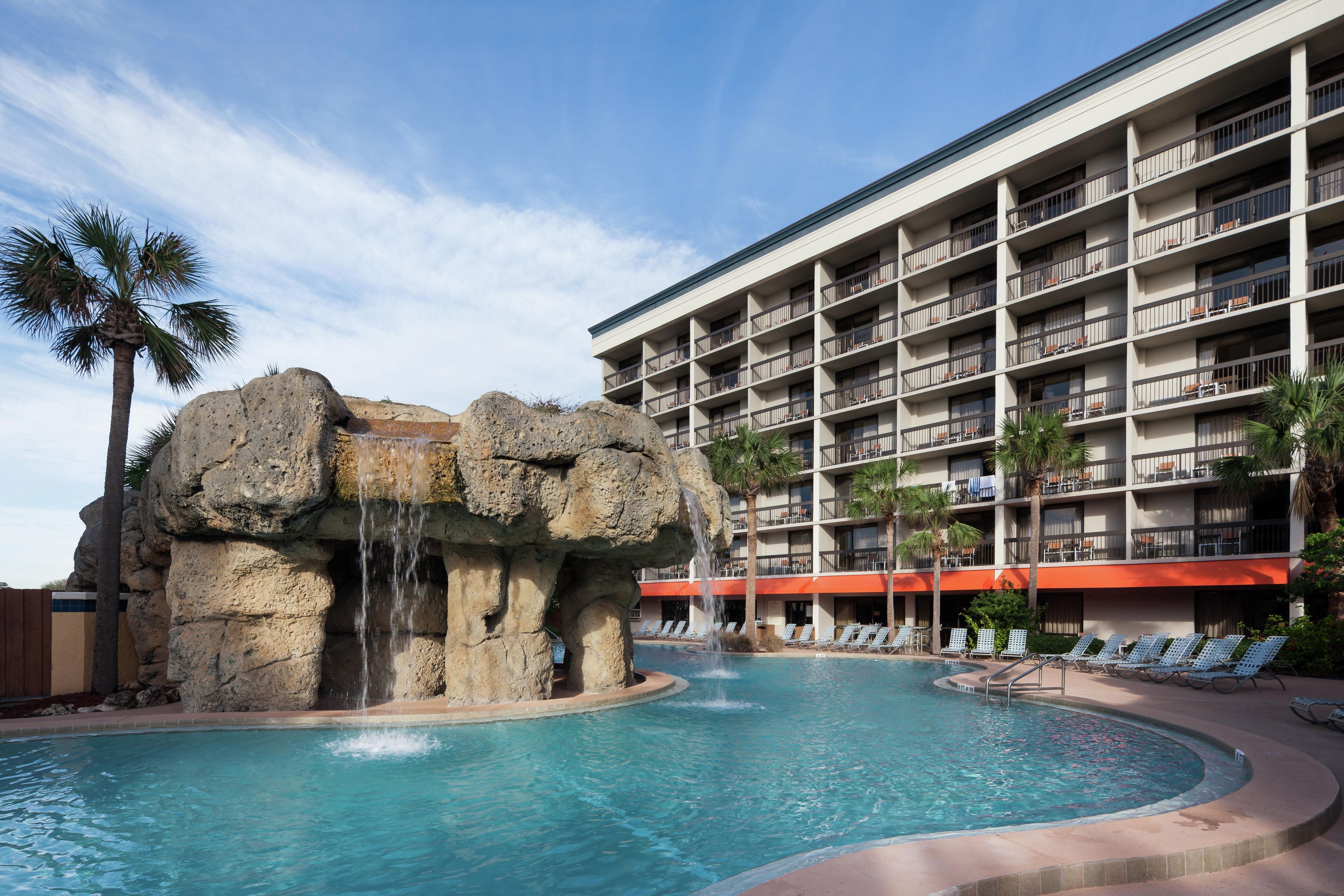 Ein Hotel mit Pool, Felsenformation mit Wasserfall, mehreren Balkonen und Palmen.
