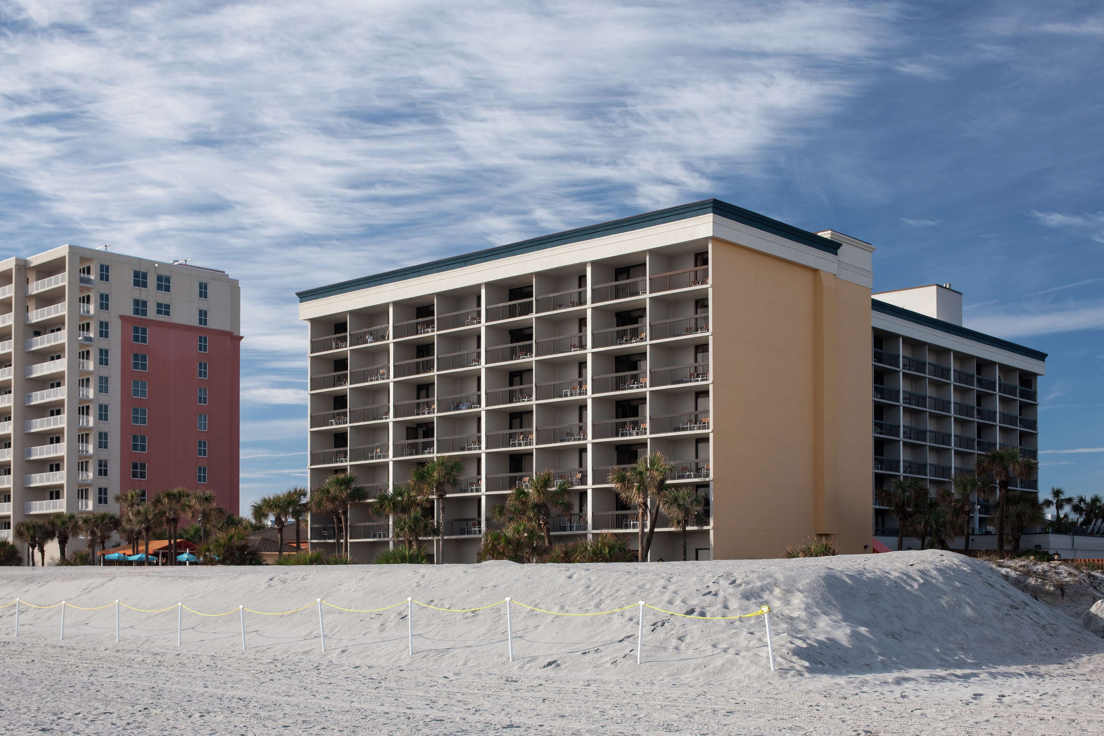 A multi-story hotel with a flat facade, multiple balconies, and a sandy foreground with a chain-link fence.