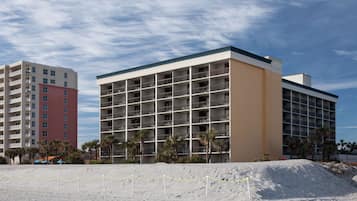 A multi-story hotel with a flat facade, multiple balconies, and a sandy foreground with a chain-link fence.