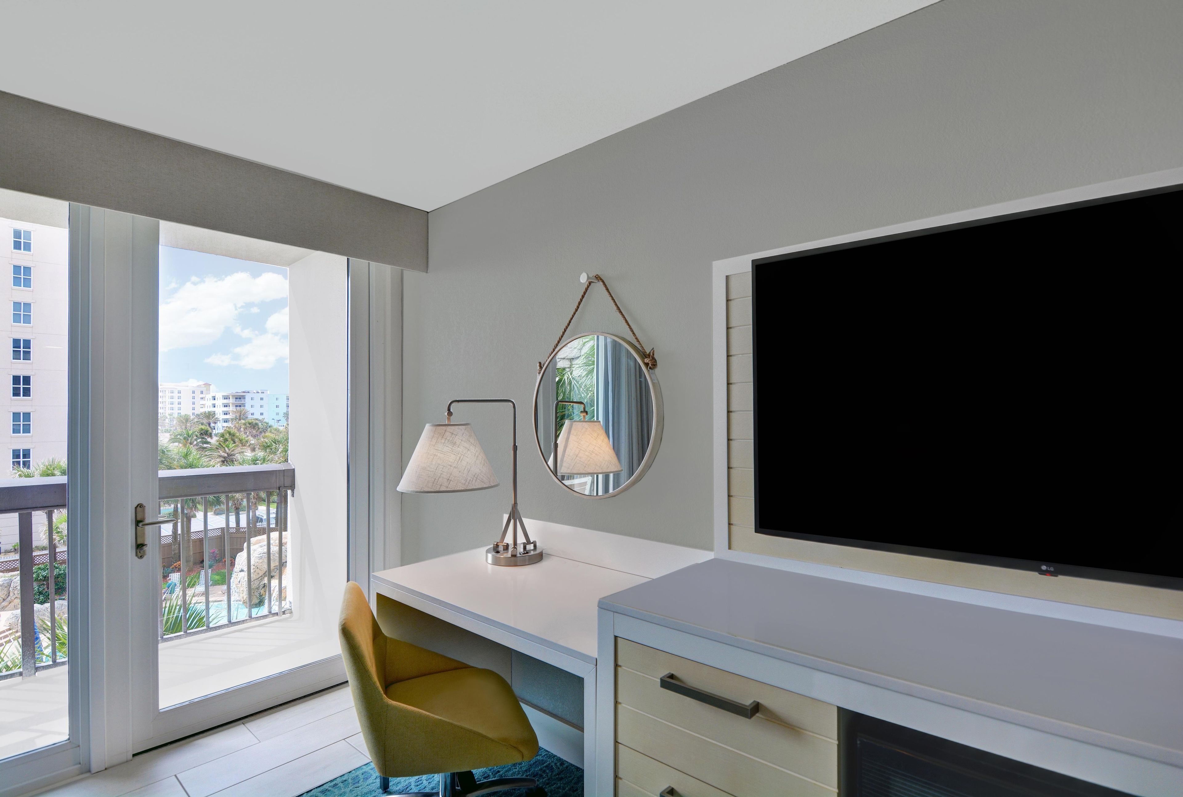 Une chambre d’hôtel avec un balcon, un bureau, une chaise, une télévision et un miroir.