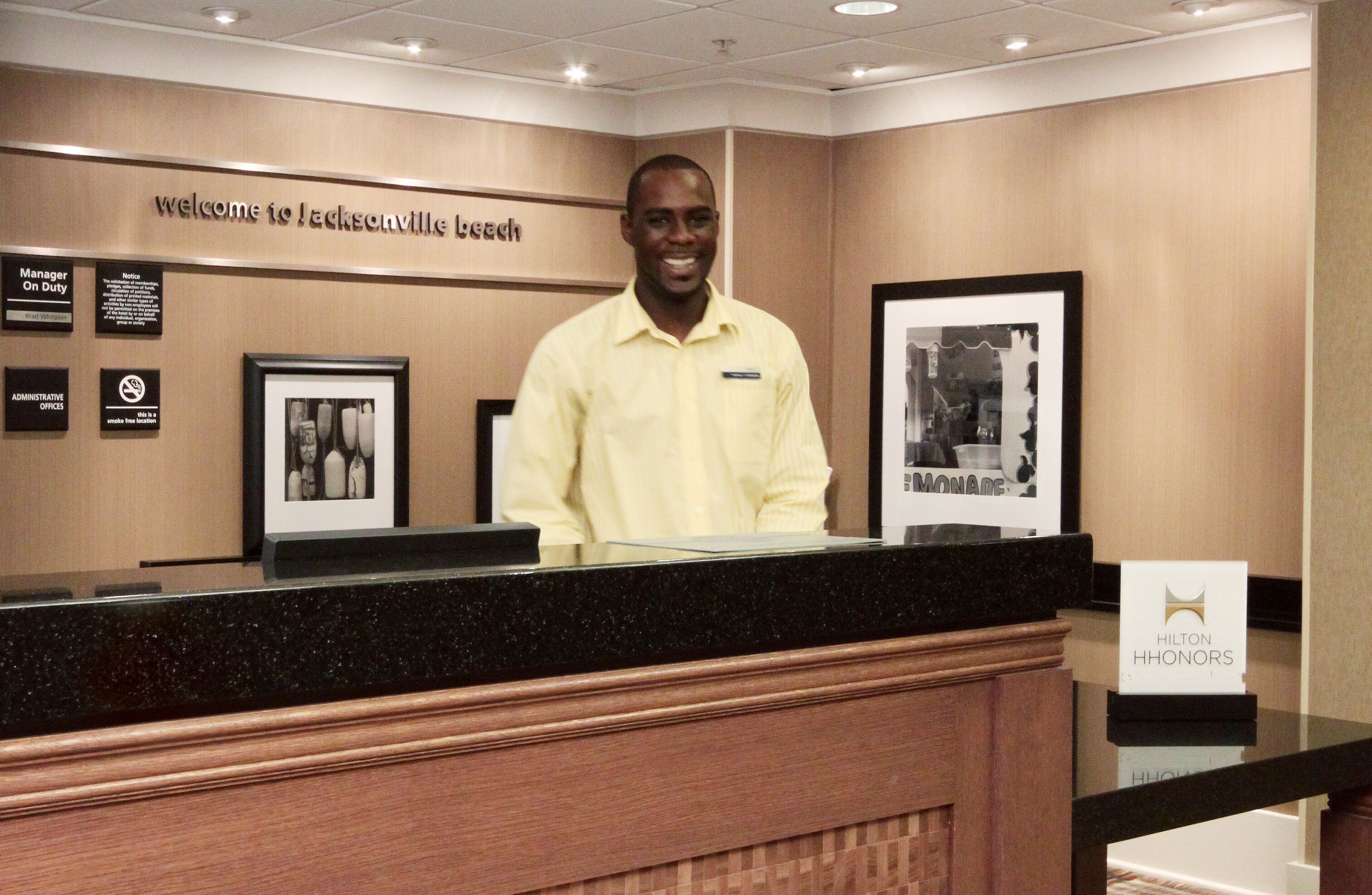 Un comptoir d’accueil d’hôtel avec une pancarte « Bienvenue à Jacksonville Beach », un tableau encadré et une pancarte « Hilton Honors ».