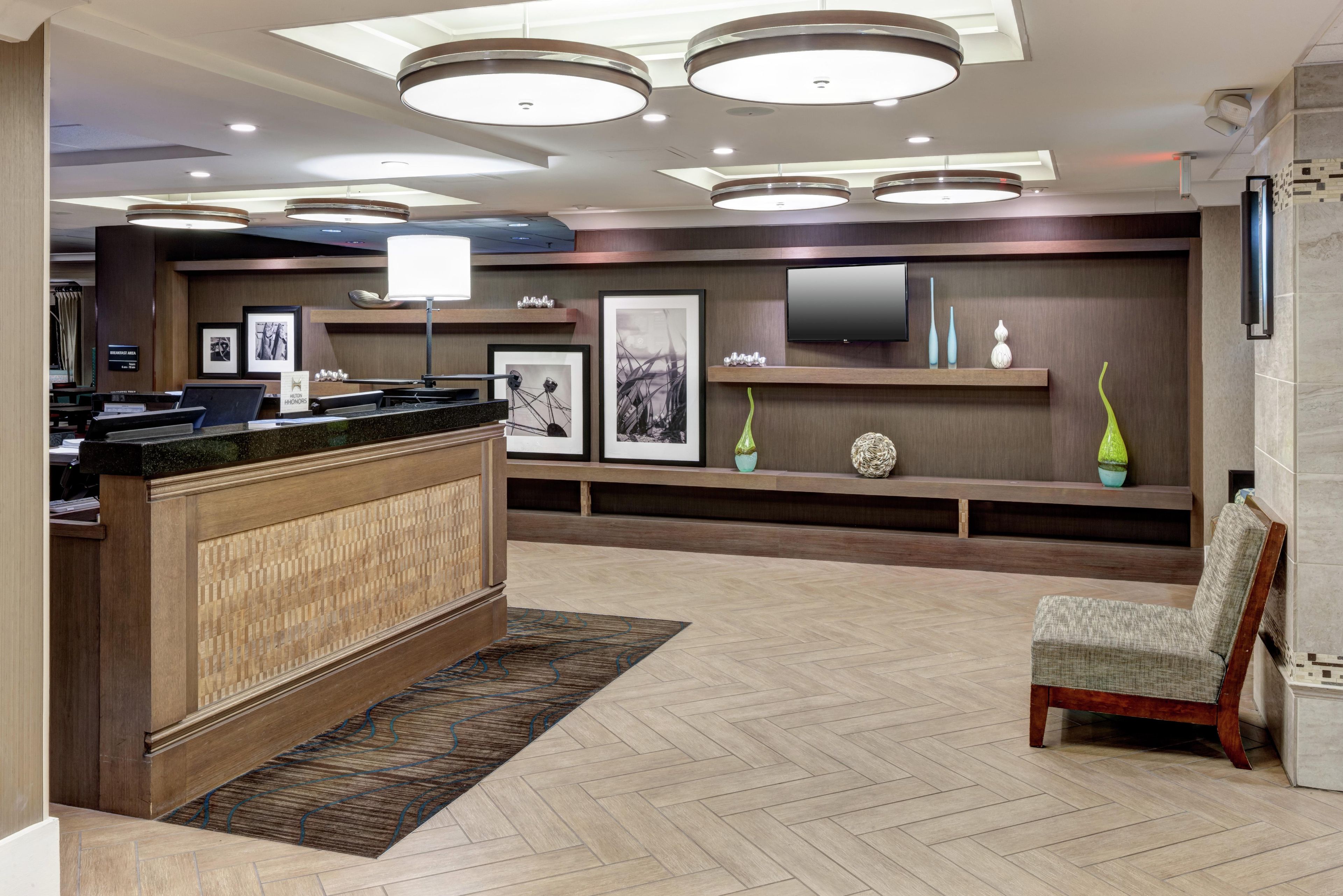 A reception area with a wooden reception desk, a chair, a flat-screen TV mounted on the wall, framed artwork, and a herringbone-patterned floor.