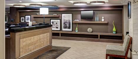 A reception area with a wooden reception desk, a chair, a flat-screen TV mounted on the wall, framed artwork, and a herringbone-patterned floor.