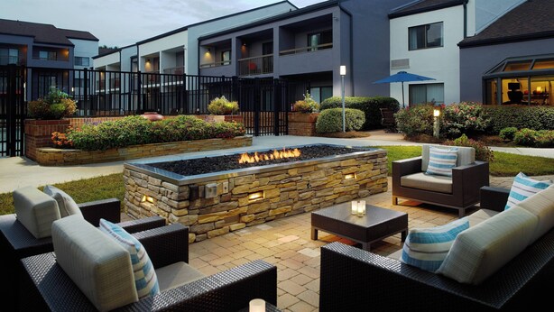 An outdoor patio area with stone fire pit, wicker furniture, and a view of the hotel buildings. - Courtyard by Marriott Raleigh Midtown (Raleigh)