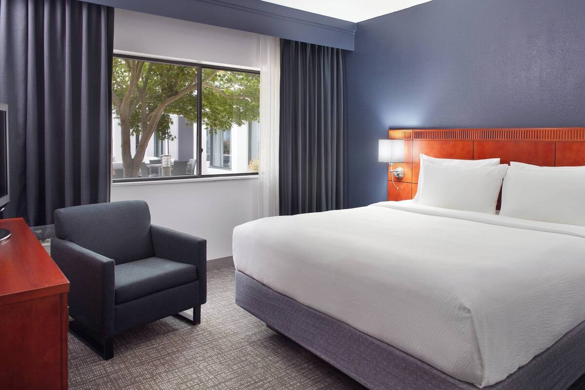 A hotel room with a large bed, a desk, a chair, a window with a view of trees, and a wall with a textured pattern.