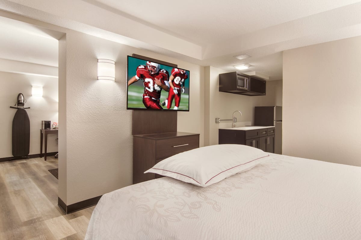 A hotel room with a large bed, a flat-screen TV mounted on the wall, a small desk with a lamp, and a kitchenette area.