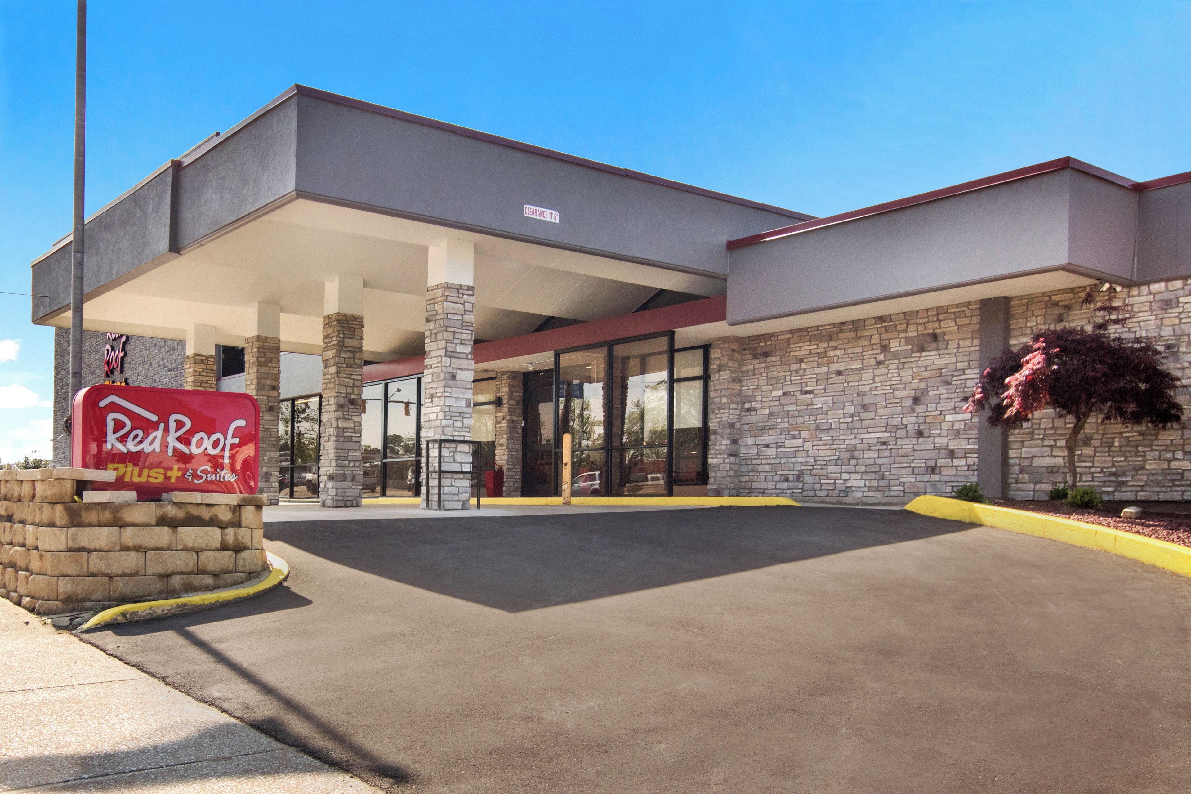 A Red Roof Plus Suites hotel with a stone facade, a covered parking area, and a clear blue sky.