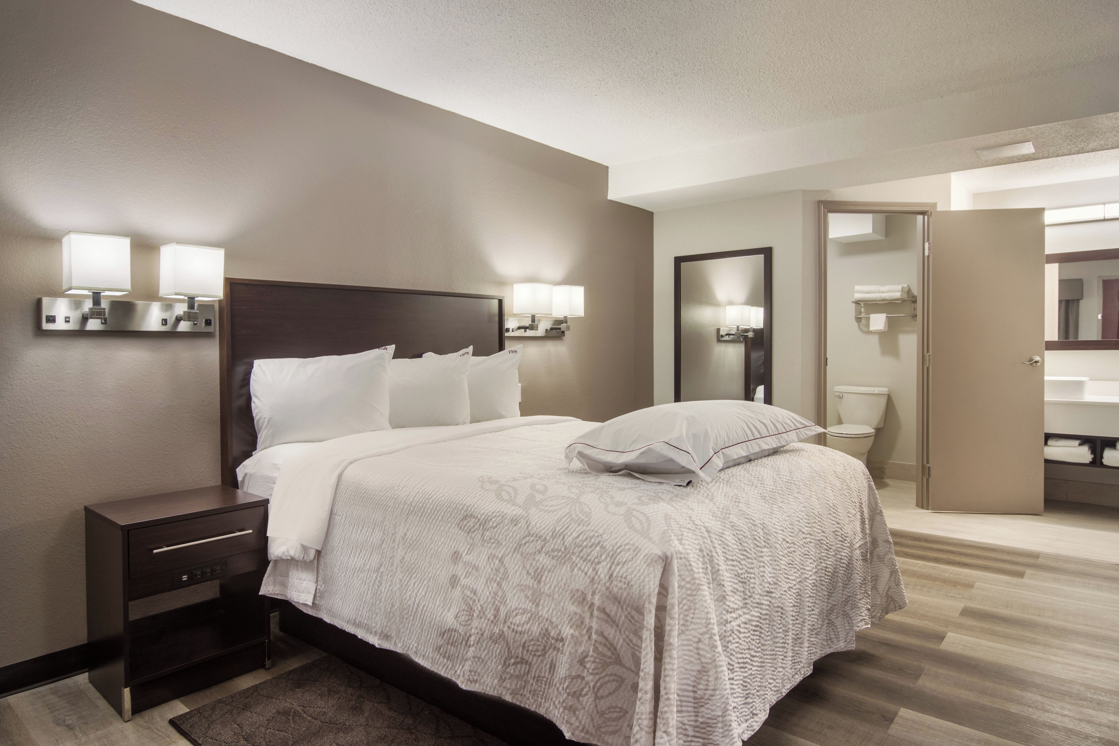Une chambre d’hôtel avec un lit, des tables de chevet, un miroir et une salle de bain visible à travers une porte ouverte.