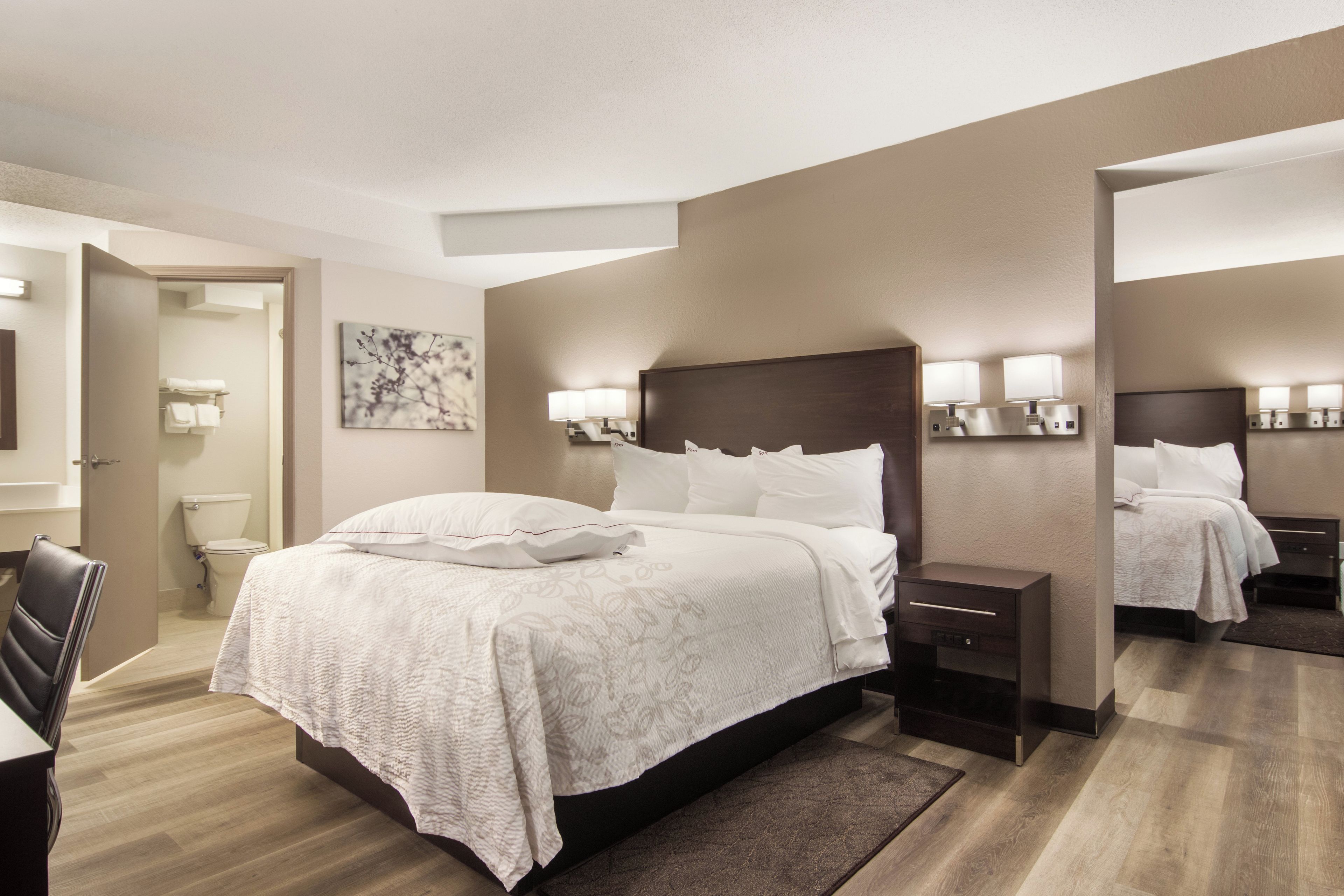 A modern hotel room with a large bed, bedside tables, a desk, and a bathroom visible through an open door.