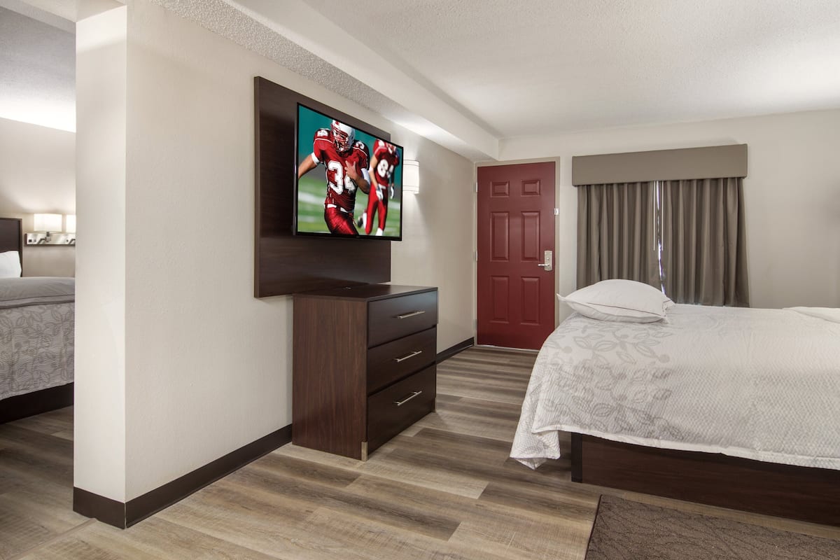 A hotel room with a large flat-screen TV mounted on the wall, a wooden dresser, a bed with a patterned bedspread, and a red door.