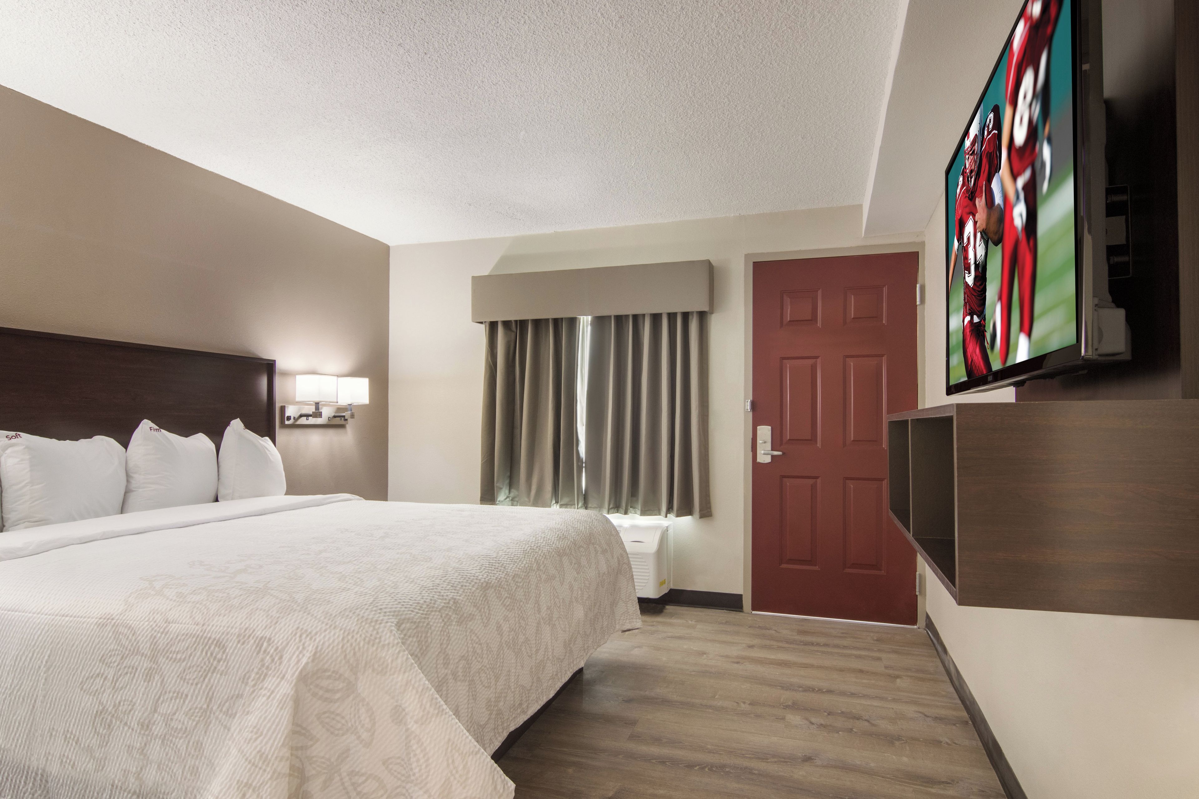 Ein Hotelzimmer mit einem großen Bett, einem an der Wand befestigten Fernseher, einer Tür und einem mit Vorhängen versehenen Fenster.
