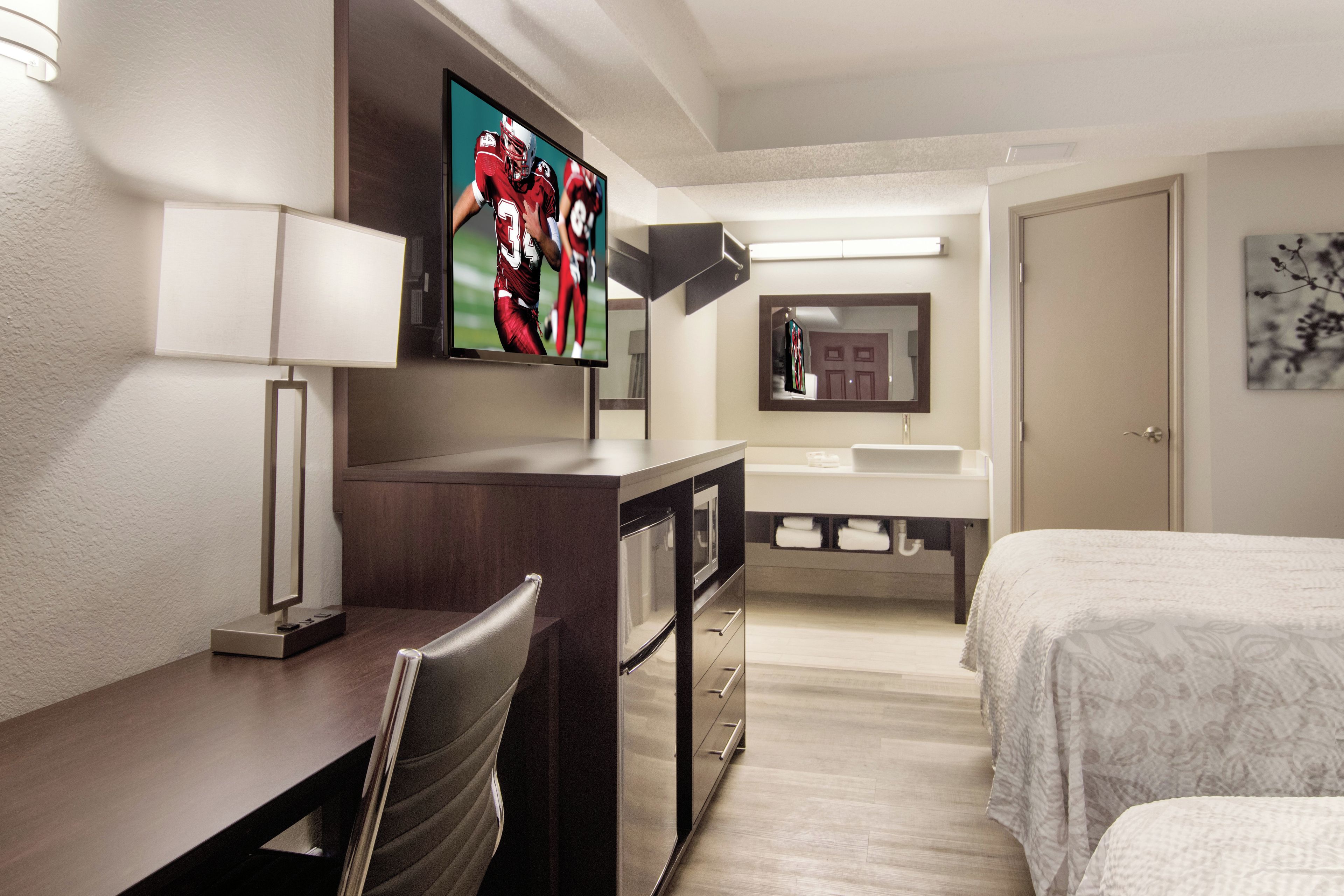 A hotel room with a bed, a desk with a chair, a television mounted on the wall, a bathroom with a sink and mirror, and a door.