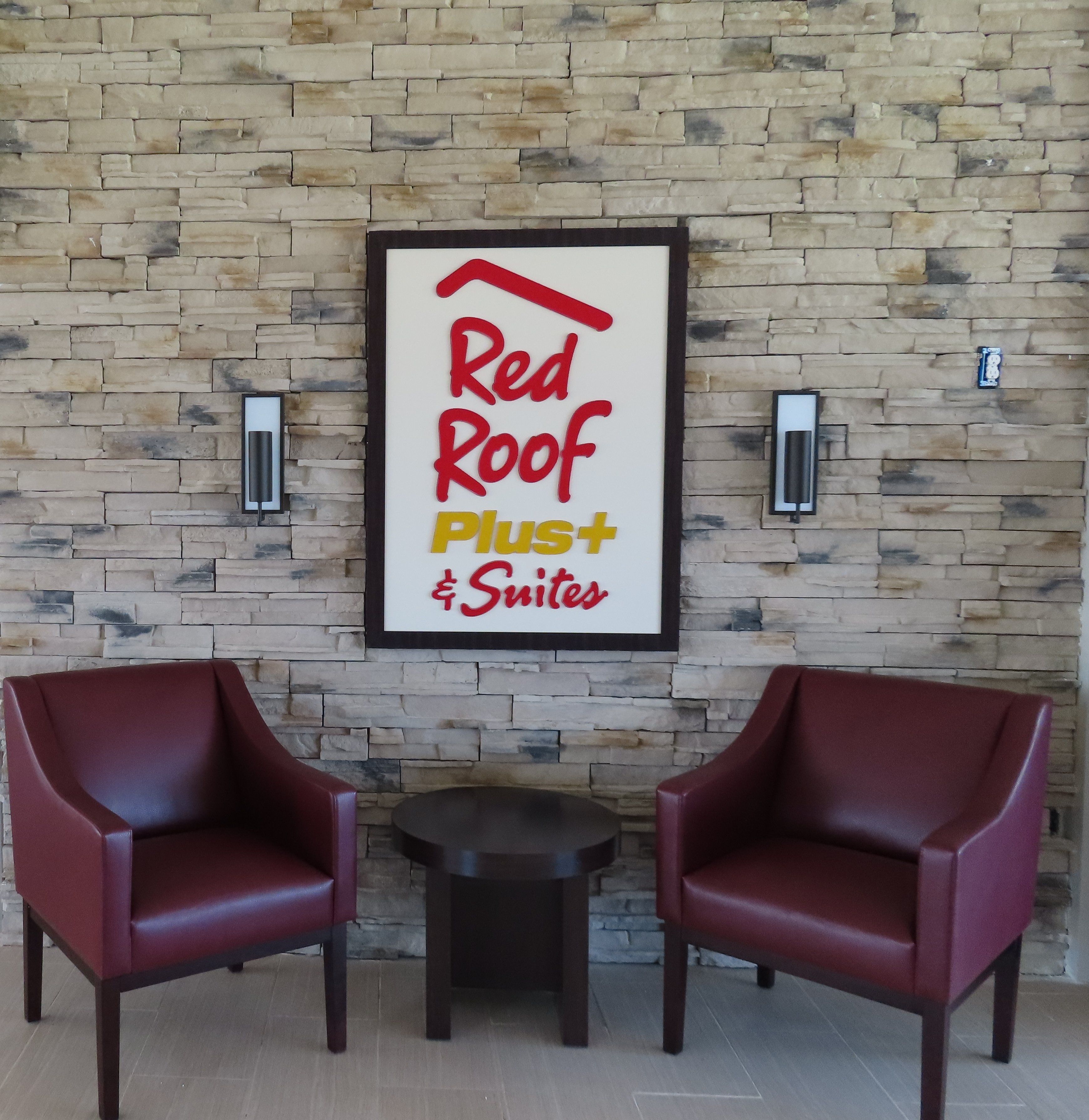 Une enseigne encadrée portant l’inscription  « Red Roof Plus Suites », fixée à un mur de pierre, avec deux chaises rouges et une petite table devant.