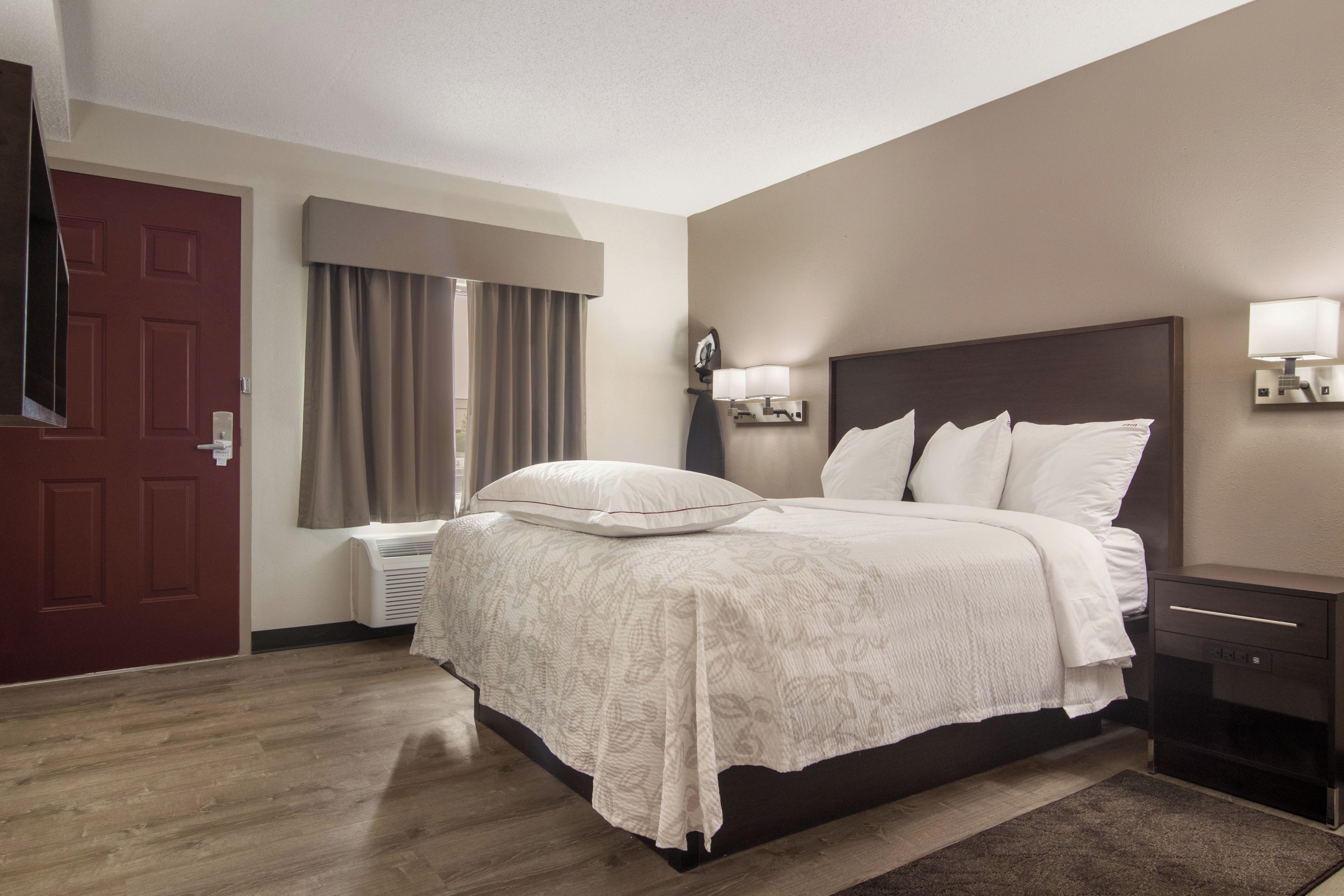 A hotel room with a bed, a nightstand, a lamp, a window with curtains, and a door.