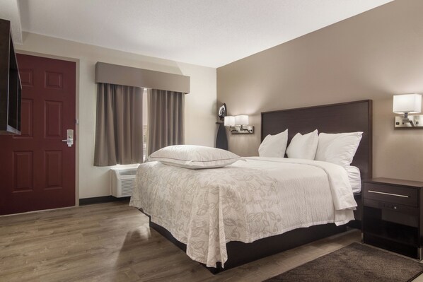 A hotel room with a bed, a nightstand, a lamp, a window with curtains, and a door. - Red Roof Inn PLUS+ & Suites Erie (Erie)