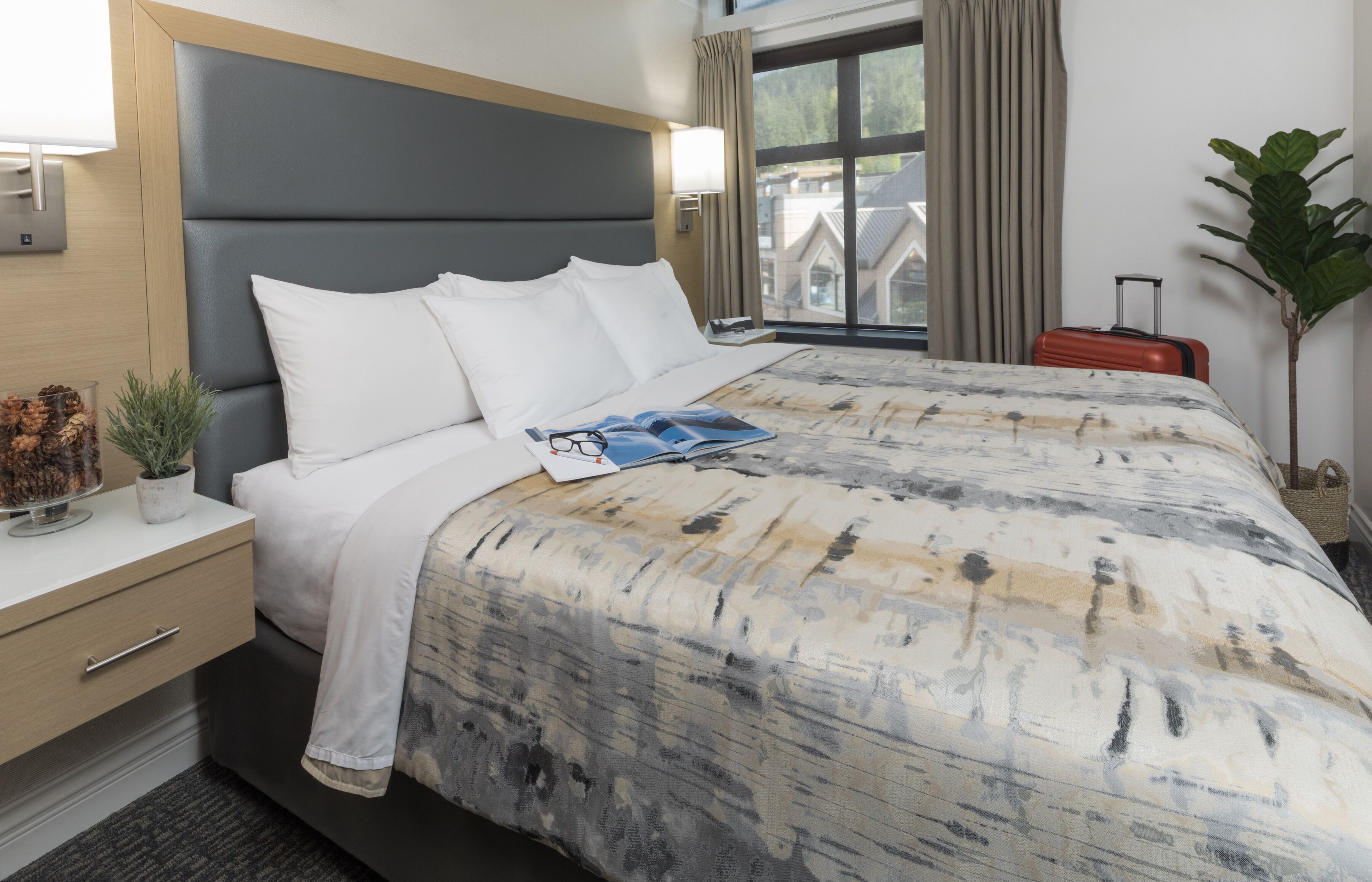 Habitación de hotel con cama, mesita de noche con lámpara, ventana con cortinas y maleta.