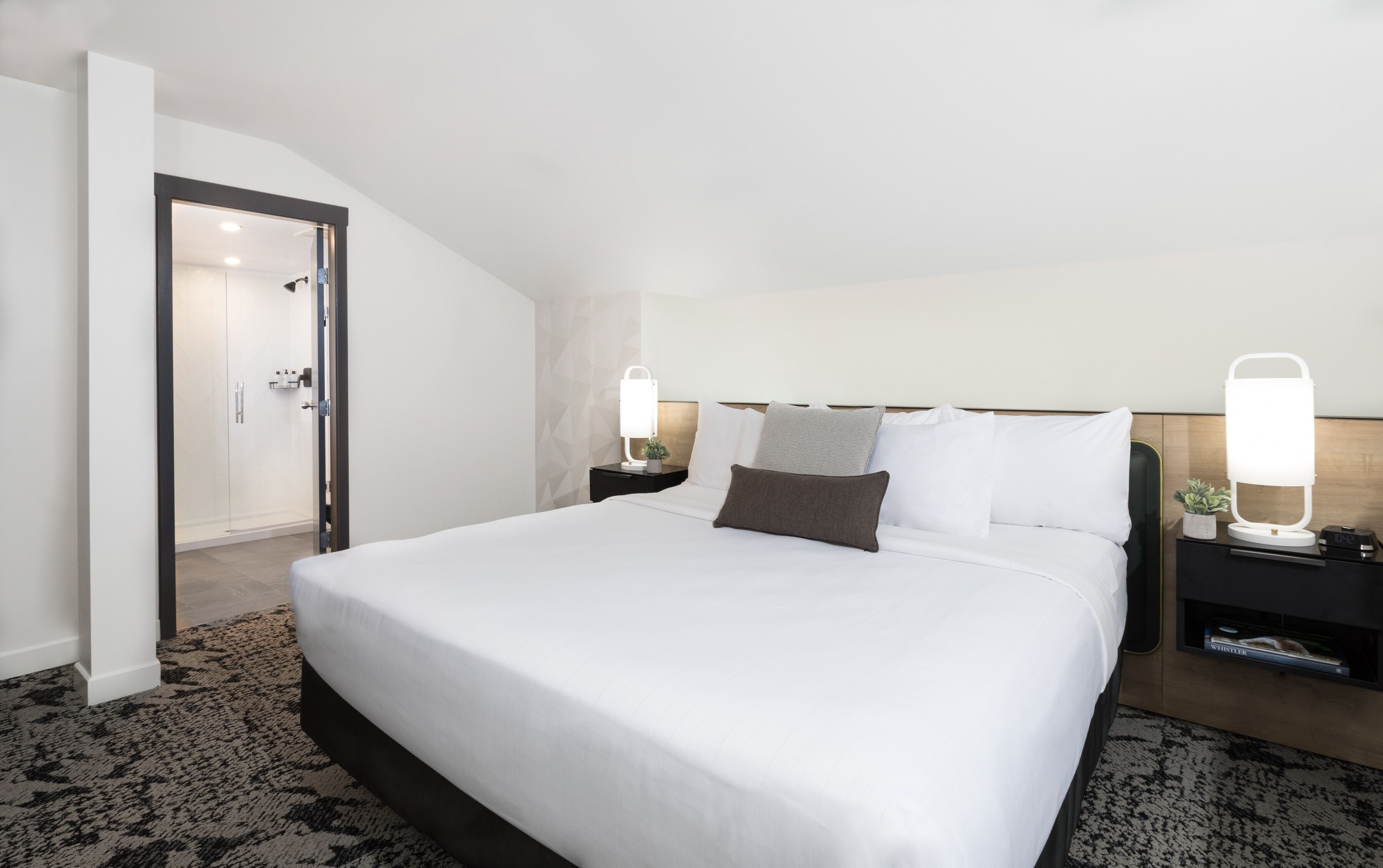 A hotel room with a large bed, two bedside tables with lamps, a door leading to a bathroom, and a small potted plant on one of the tables.