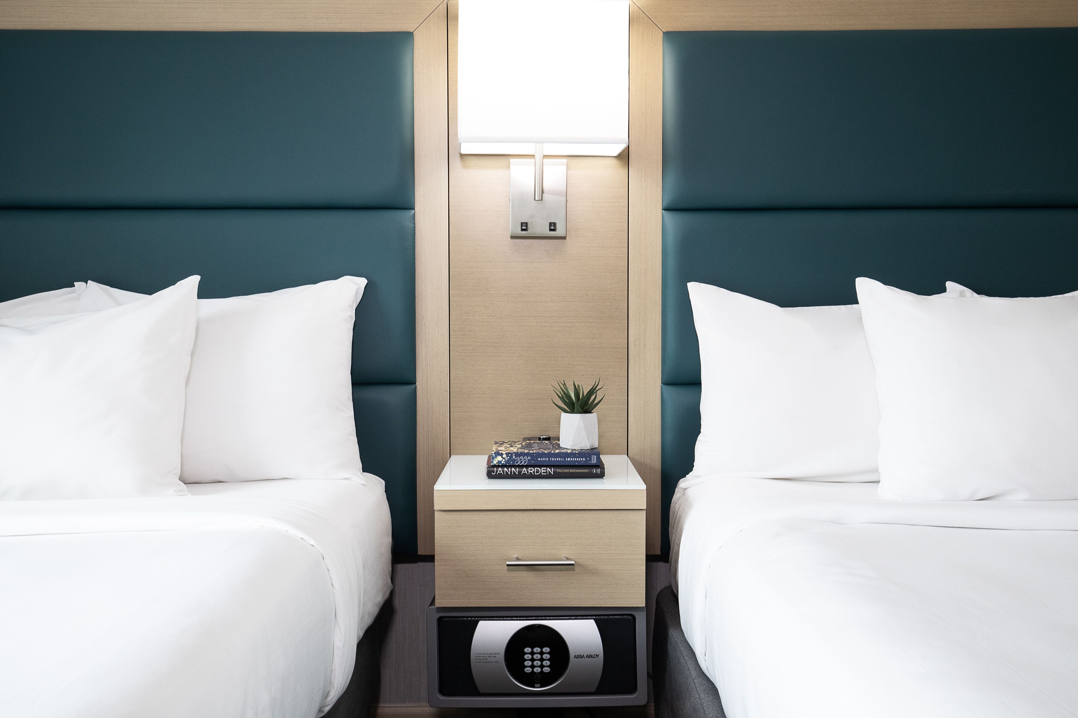 A hotel room with two beds, a bedside table with a lamp, a small plant, and a book titled "JANN ARDEN".