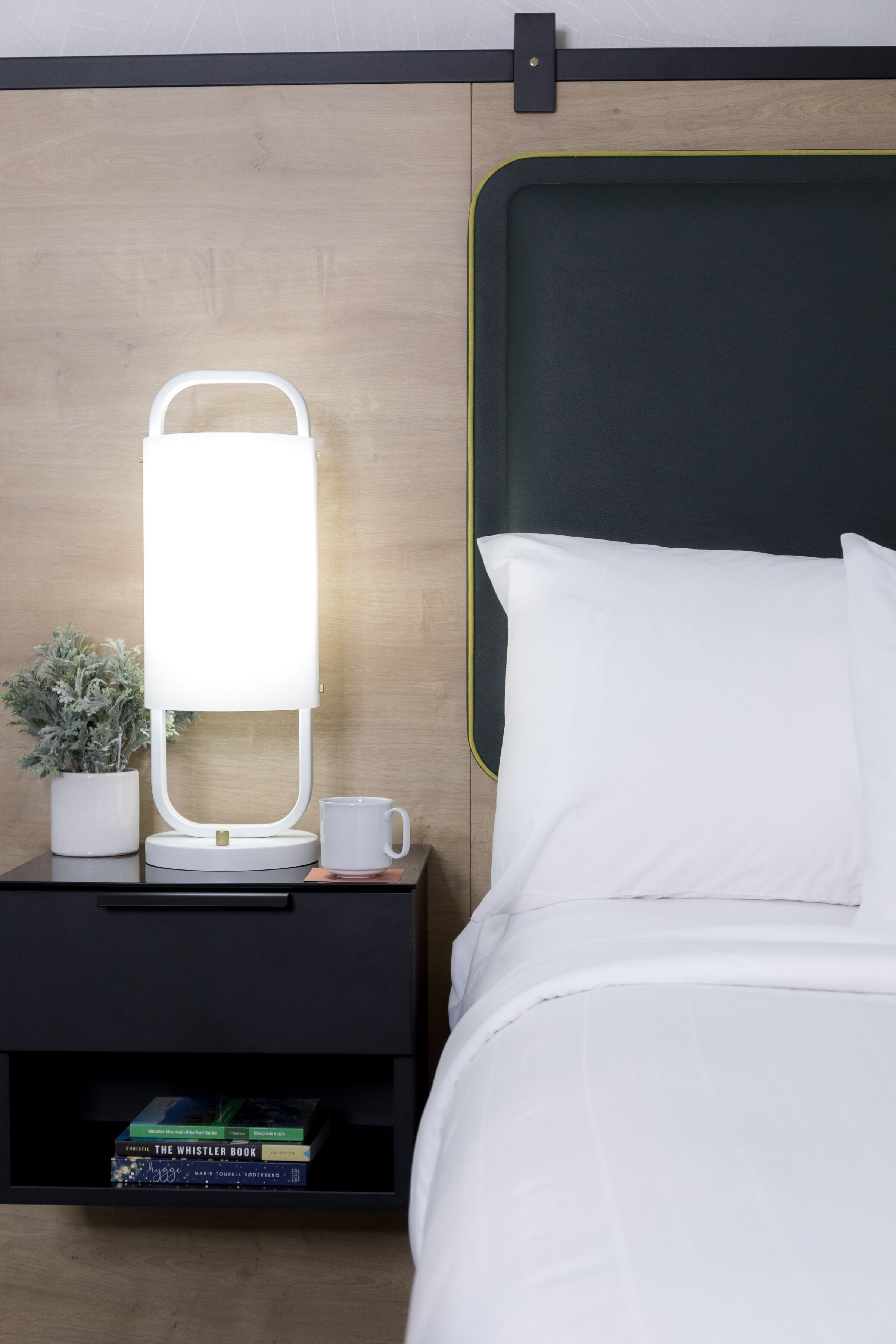 Un lit bien fait, avec du linge de lit blanc, une table de chevet surmontée d’une lampe blanche, une petite plante en pot et une tasse blanche. Une étagère se trouve sous la table, où sont posés quelques livres.