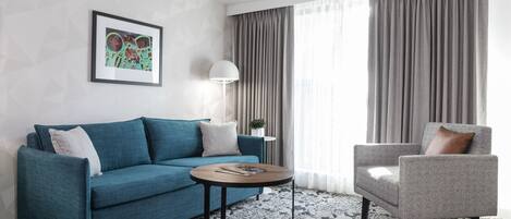 Una sala de estar moderna con un sofá verde azulado, un sillón gris, una mesa redonda de centro y un cuadro en la pared.