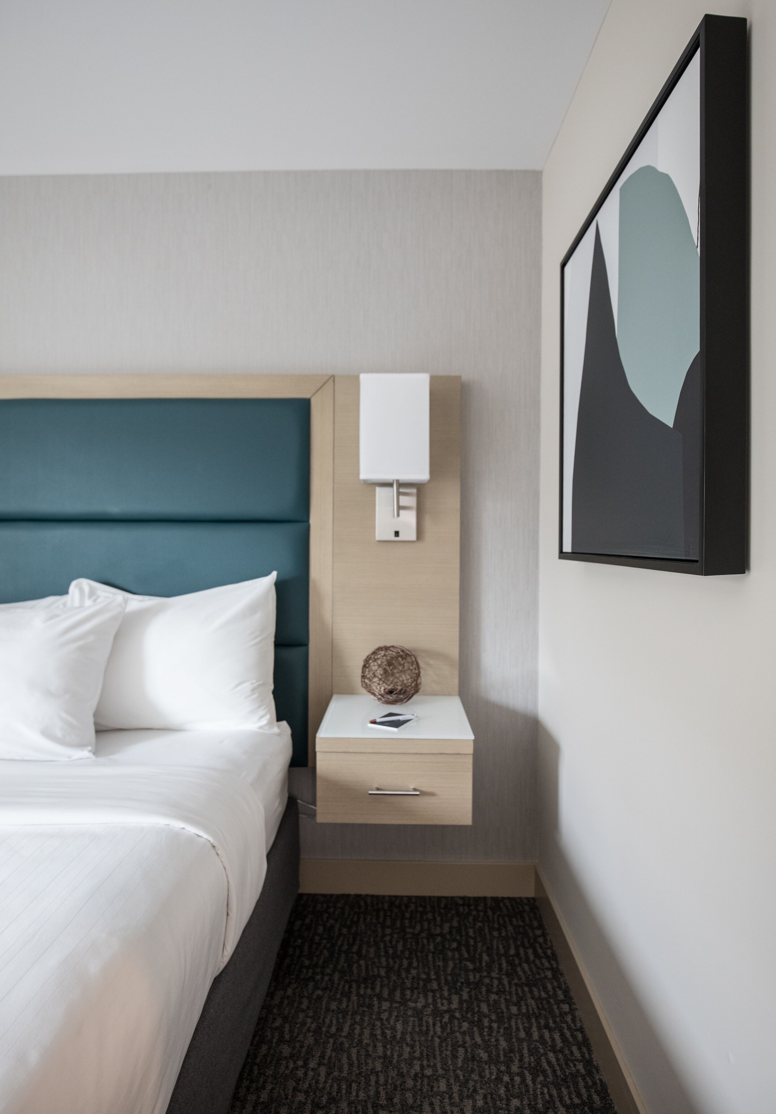 A hotel room with a bed, a bedside table with a lamp, a framed picture on the wall, and a carpeted floor.