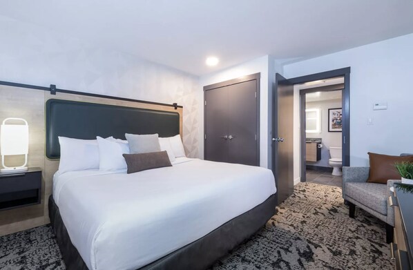 Una habitación de hotel moderna con una cama grande, una mesita de noche, un escritorio, una silla y una puerta que da a un baño.