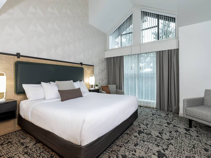 A modern hotel room with a large bed, a nightstand with a lamp, a sofa, and a large window with curtains.