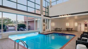 Una piscina interior con paredes de vidrio transparente, suelo de baldosas y un área de estar con mesas y sillas.
