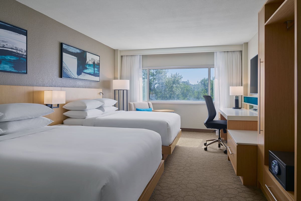A hotel room with two beds, a desk, a chair, a TV, and a window with a view of trees.