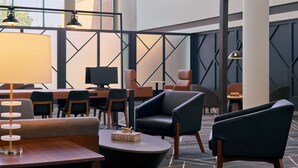 Un moderno vestíbulo de hotel con zona de estar, mesas y un amplio ventanal que proporciona luz natural.