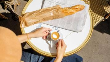 Una persona sentada a una mesa redonda con un periódico, una baguette y una taza de café.
