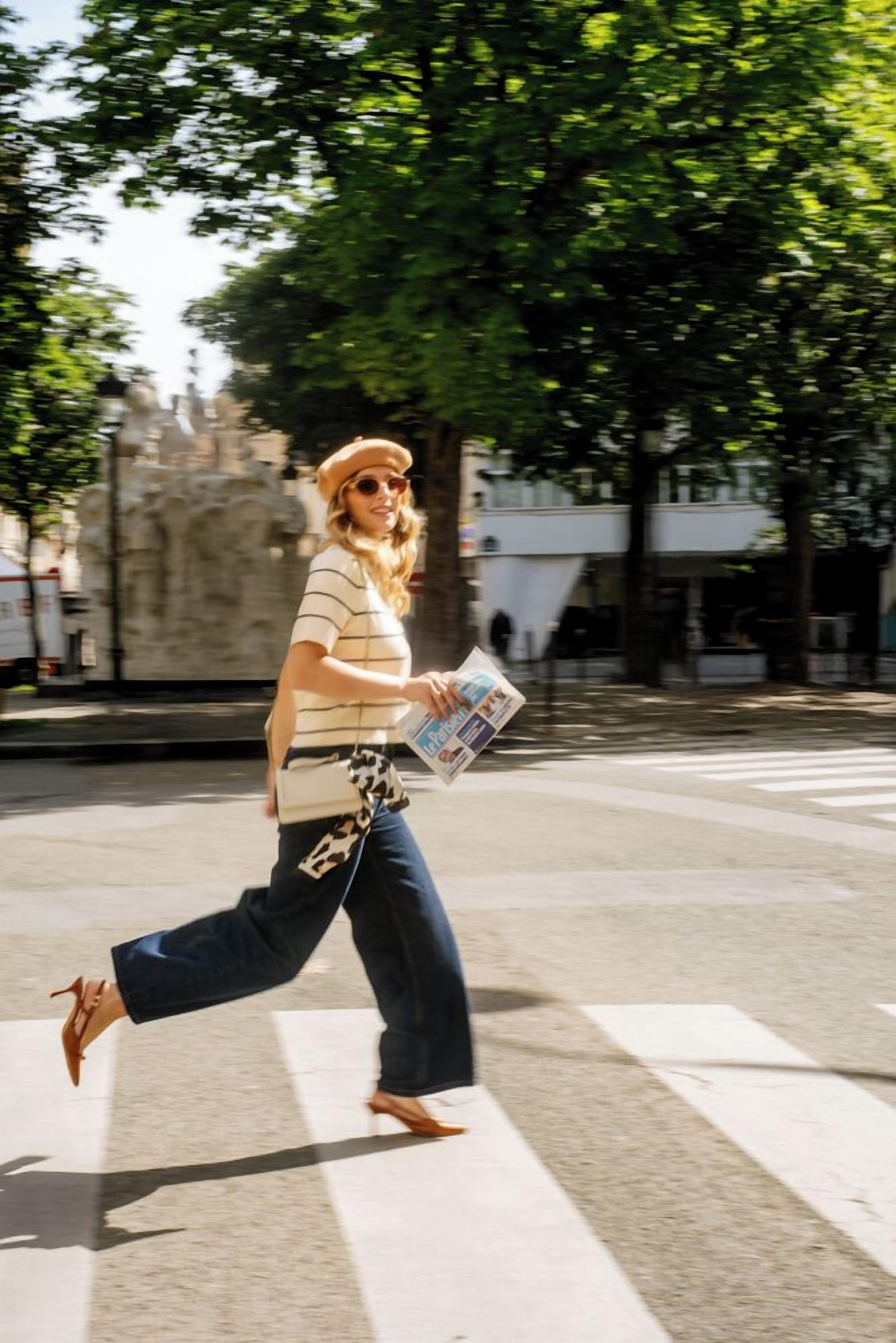 Une personne traversant une rue, tenant un journal à la main, avec des arbres et un bâtiment en arrière-plan.