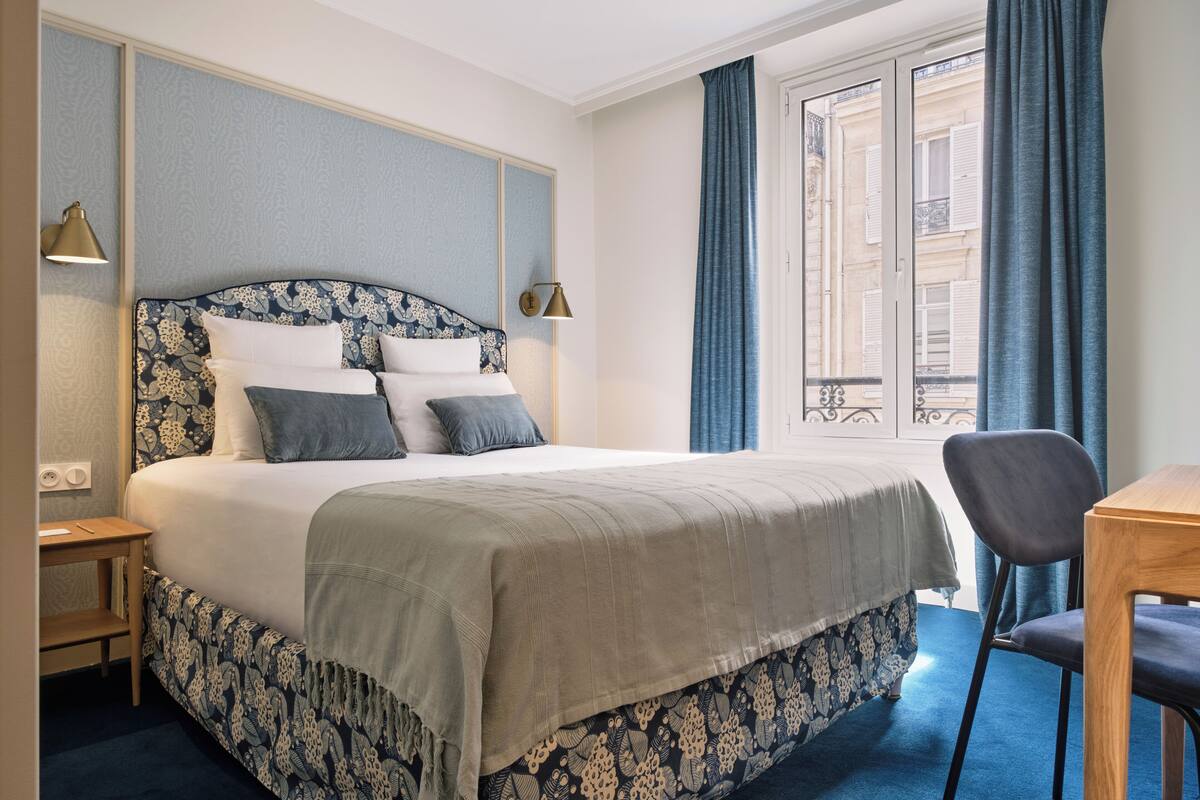 A hotel room with a large bed, a wooden side table, a chair, and a view of the city through the window.