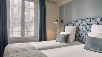 Habitació d'hotel amb un llit gran, dos almoinos, una tauleta de nit, una làmpada i una finestra amb cortines.