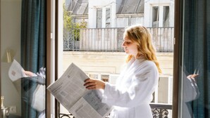 A person in a white robe standing by a window with a newspaper, a mirror, and a view of buildings outside.