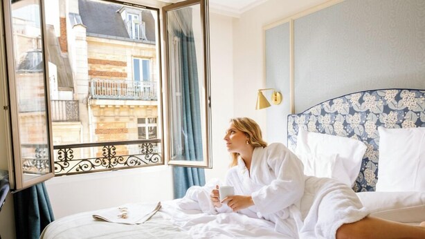 A hotel room with a bed, a window with a view of buildings, and a bedside lamp.