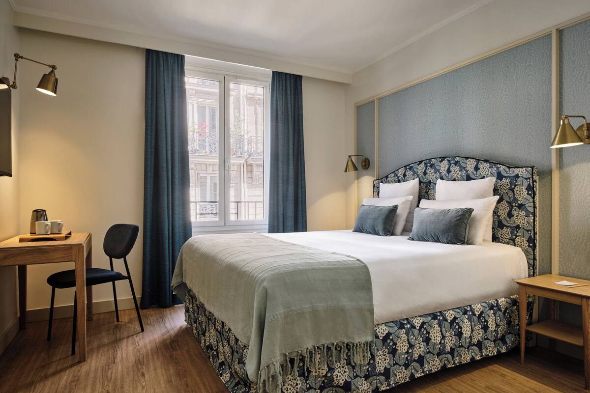 A hotel room with a large bed, a desk, a chair, and a window with curtains.